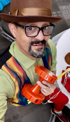 A smiling man wearing a hat, glasses, and colourful vest holds a juggling toy. His rainbow socks are just visible.