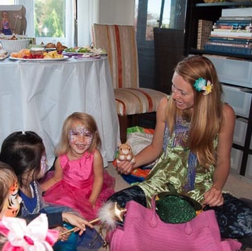 A magician sits on the floor next to a few little girls, holding a toy hamster and performing a magic show