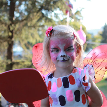 A little girl wearing pink fairy wings looks into the mirror at her pink tiger facepaint