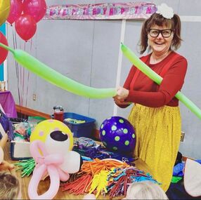 A woman twisting green balloons wears a colourful outfit. She is surrounded by balloon animals.