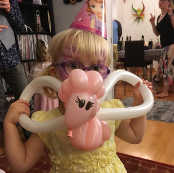 A little girl wearing glasses looks into the camera while holding a pink and white flying unicorn balloon