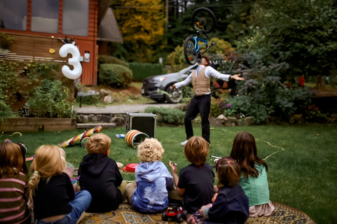 A crowd of children watch a magician balancing a bicycle on his chin at a birthday party