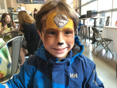 A little boy smiling with yellow, silver and brown Paw-Patrol face paint